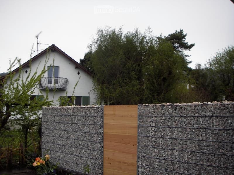 A garden area with a stone and wooden fence, featuring a white house with a balcony in the background and lush green trees.