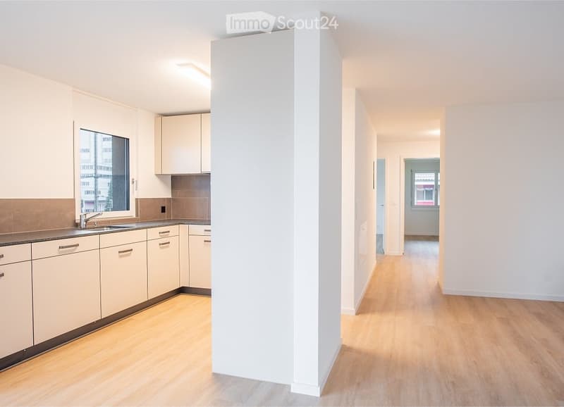 Empty room with light wooden flooring, white walls, and a small kitchen area with cabinets and a sink visible in the background.