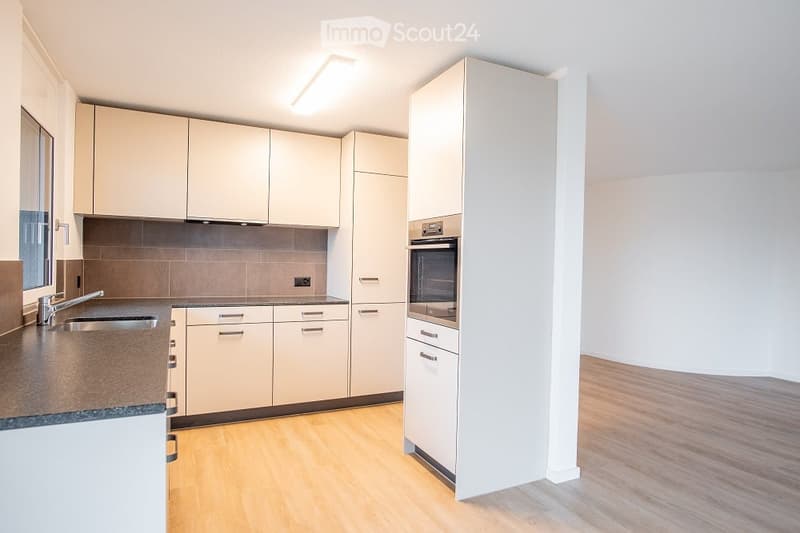 Modern kitchen with light-colored cabinets, a stainless steel sink, and an oven. The floor is light wood, and the walls are white with a tiled backsplash.
