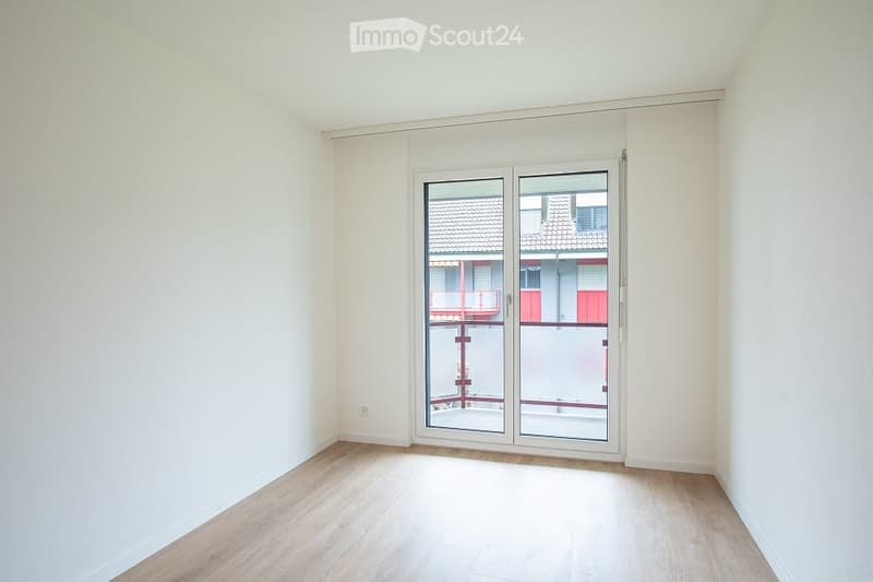 Empty room with light wooden flooring and white walls, featuring a large glass sliding door leading to a balcony.