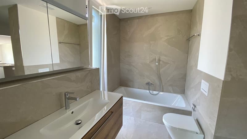 Modern bathroom with a large mirror, wooden cabinet, sink, bathtub, and toilet. The walls are tiled in a neutral color, and the floor is also tiled.