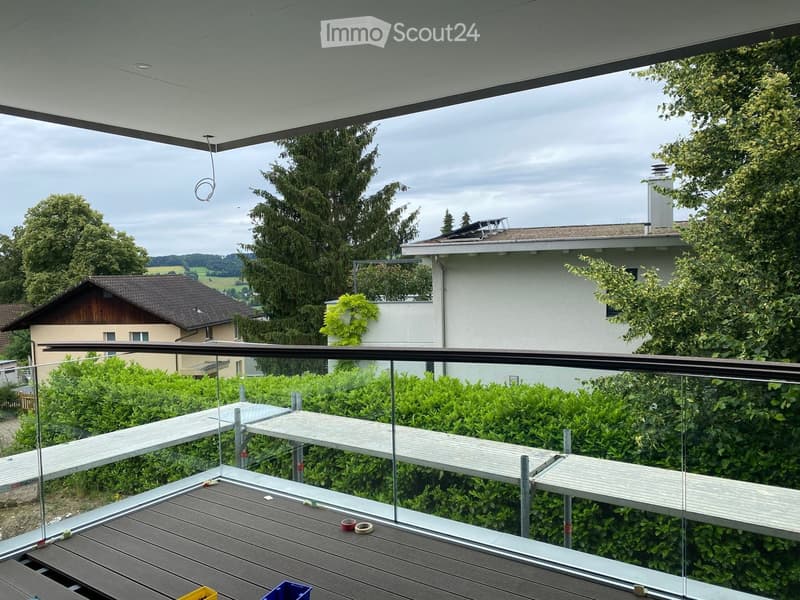 Balcony with glass railing, wooden decking, and surrounding greenery, overlooking a residential area with houses and trees.