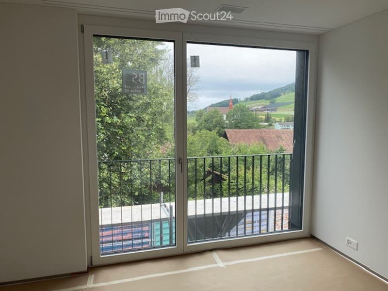 Empty room with large sliding glass doors opening to a balcony, showing a scenic view of greenery and a town with a church tower.