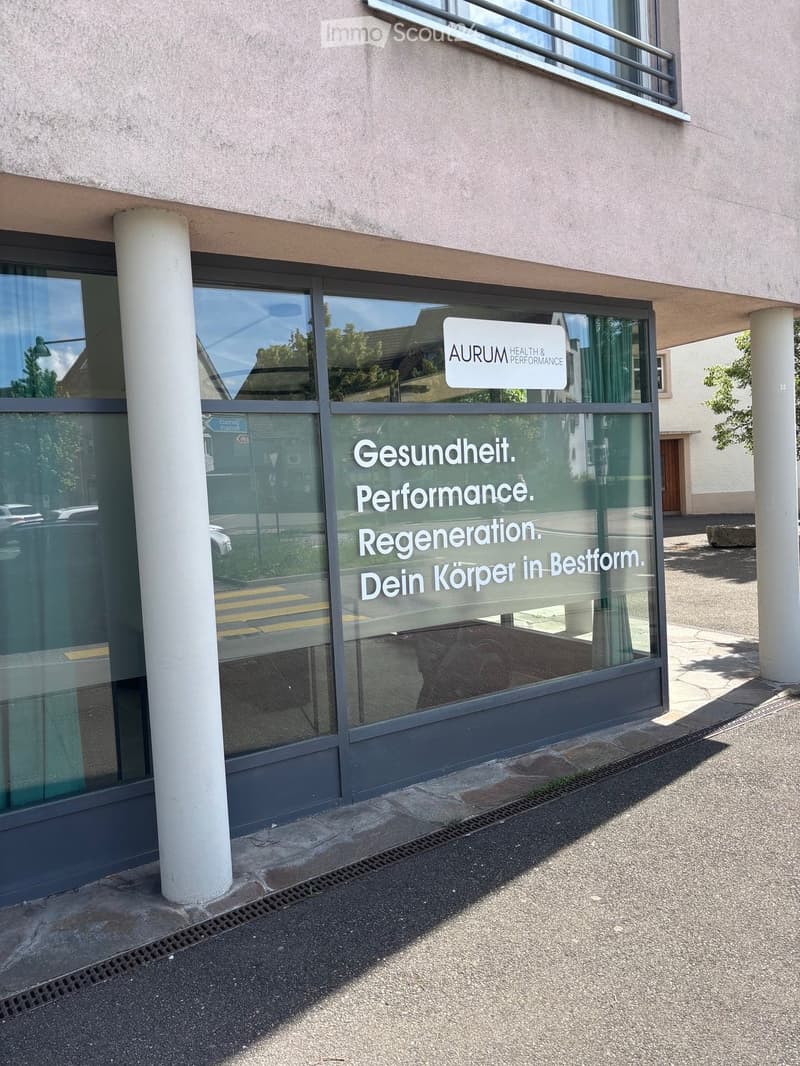 Modern building facade with large glass windows and a sign reading "AURUM HEALTH & PERFORMANCE" and additional text in German on the glass.