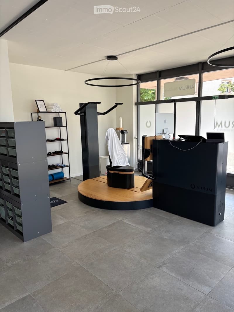 Modern, minimalist interior with a round wooden platform, a black stool, a shelf with folded towels, and a reception desk with a screen.