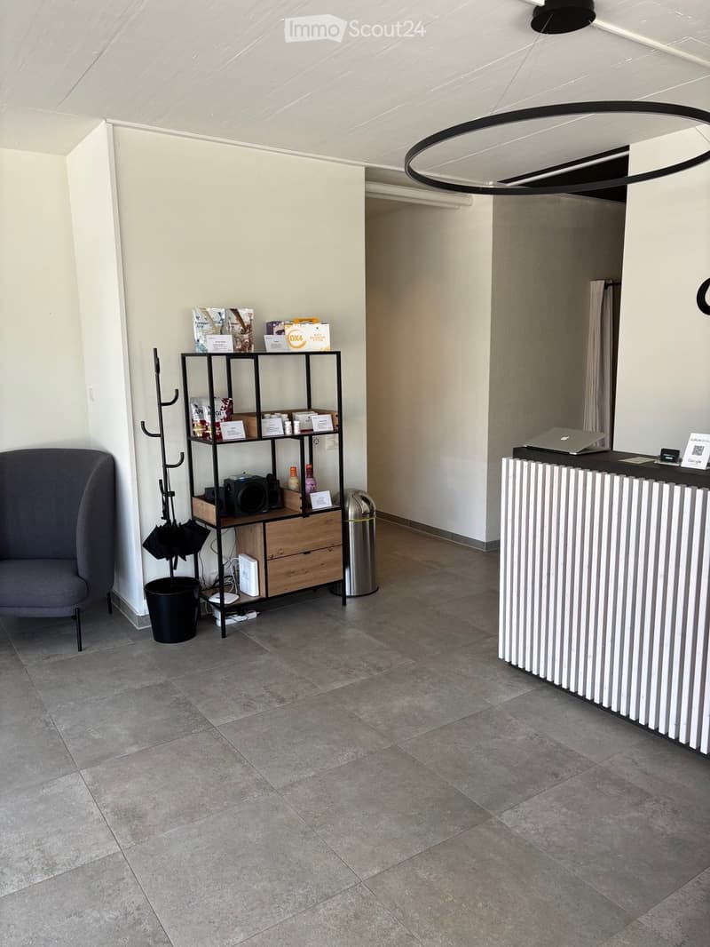 Modern reception area with a grey chair, a black metal shelf holding various items, a white radiator, a laptop on the counter, and an umbrella stand near the wall.