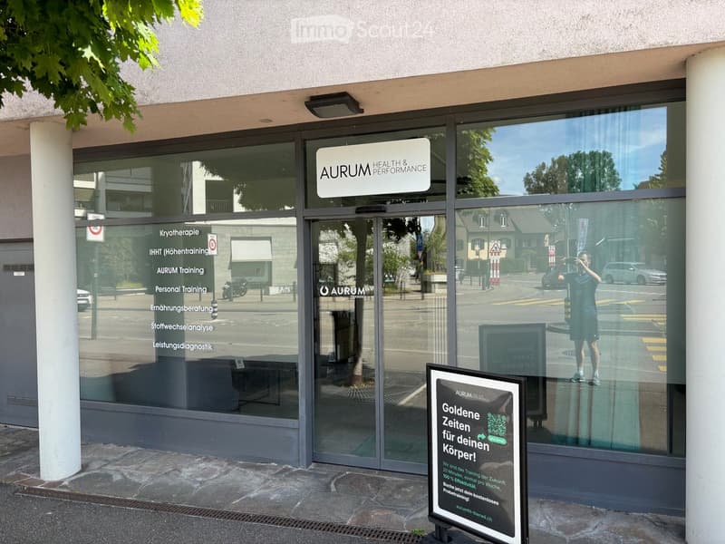 The image shows the exterior of a modern building with large glass windows and a sign reading "AURUM HEALTH & PERFORMANCE". A promotional sign in front advertises a training program. The building has a clean, contemporary design with a visible entrance an