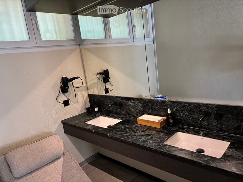 Modern bathroom with a dark marble countertop, two sinks, a large mirror, and a hair dryer mounted on the wall. The countertop features a wooden soap dish and a soap dispenser.