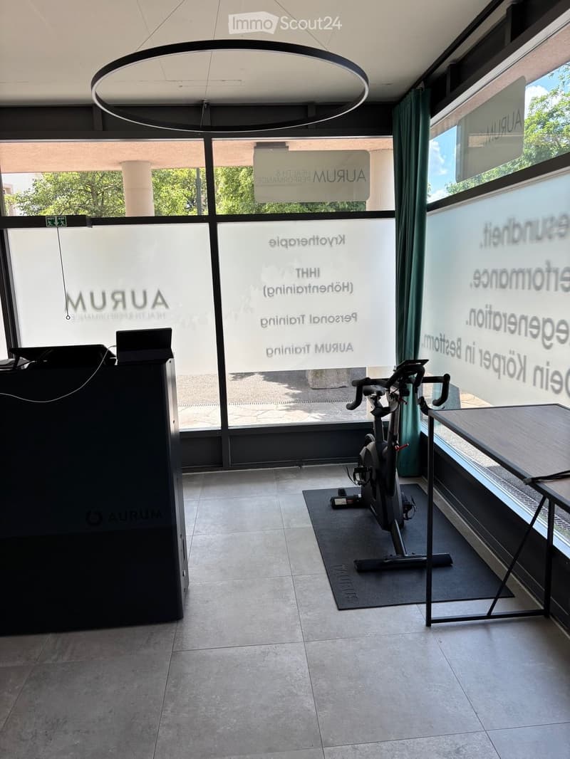 Modern, well-lit room with large windows, an exercise bike on a mat, a small table with a screen, and a sleek black cabinet.