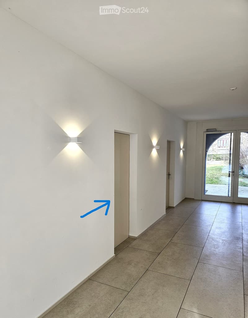 Empty hallway with white walls, tiled floor, and recessed lighting. Features a glass door leading outside and several closed doors along the walls.
