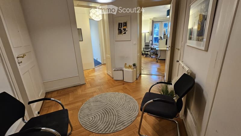 Hallway with wooden flooring, three black chairs, and a round rug. The hallway leads to another room with a desk and office chair.