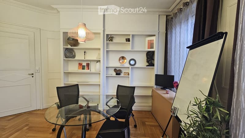 A modern, well-lit room with a glass table, two office chairs, built-in shelves, a screen, and a decorative lamp.