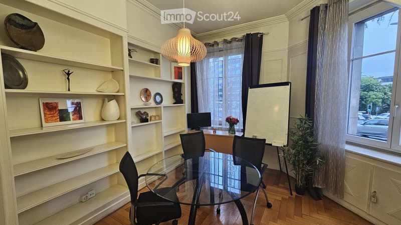 A well-lit room with a glass table and chairs, a large shelf with decorative items, a framed picture, a vase with flowers, and a screen. The room has wooden flooring and large windows with curtains.