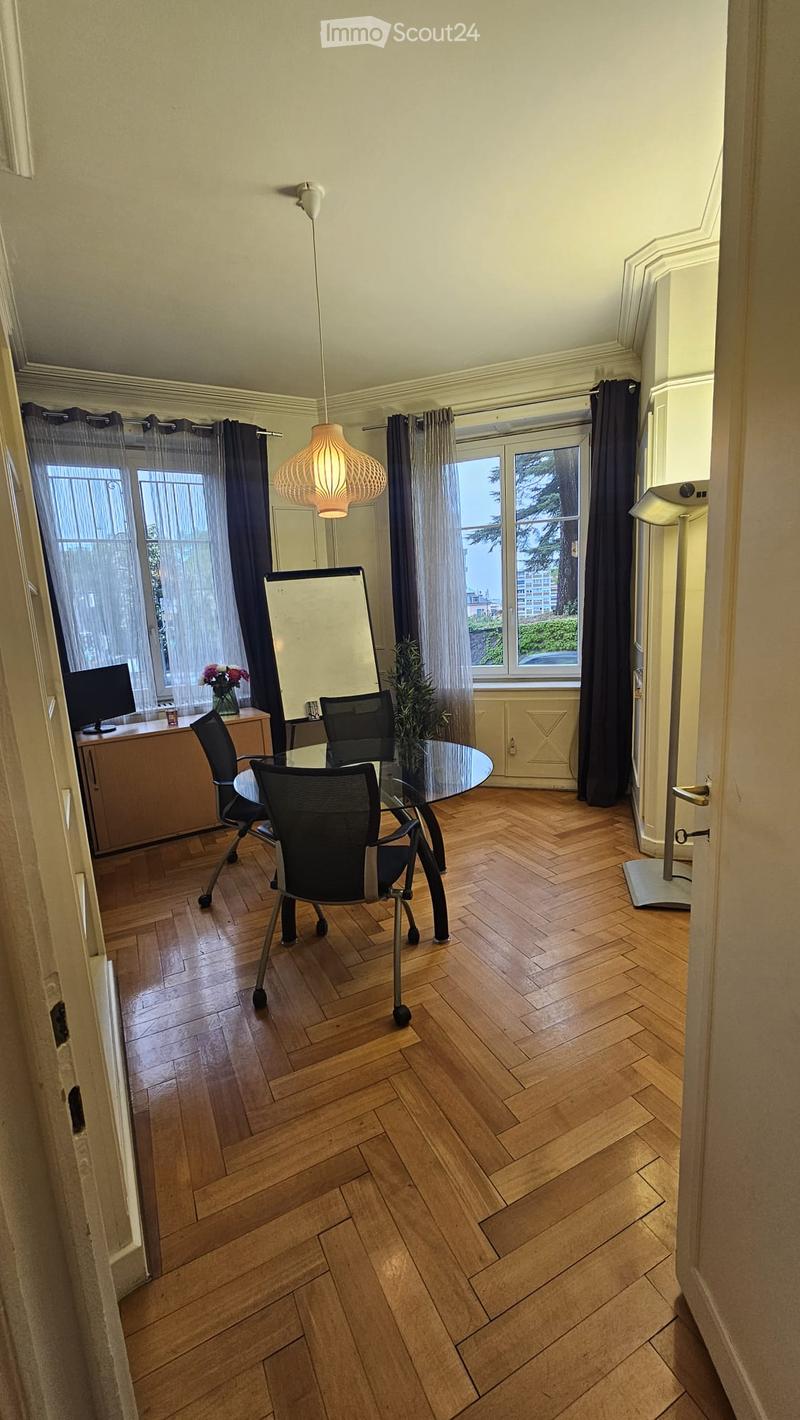 A well-lit room with wooden herringbone flooring, large windows with dark curtains, a modern pendant light, a glass table with two black chairs, a cabinet with a screen, and a small plant on the table.
