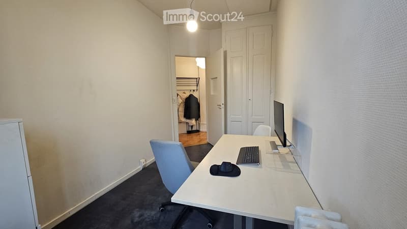 A small, furnished room with a white desk, a computer monitor, a keyboard, a mouse, a hat, and a blue office chair. The room has white walls, a hanging light fixture, and an open closet with clothes visible.
