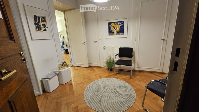 A small, well-lit hallway with wooden flooring, two chairs, a round rug, a small plant, and two picture frames on the walls.