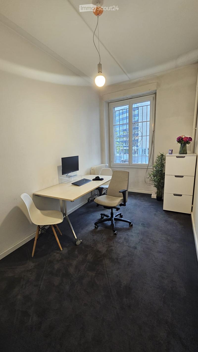 A modern, furnished room with a desk, computer, chairs, filing cabinet, and decorative plant. The room has a large window with a view of the street and is illuminated by a hanging light fixture.