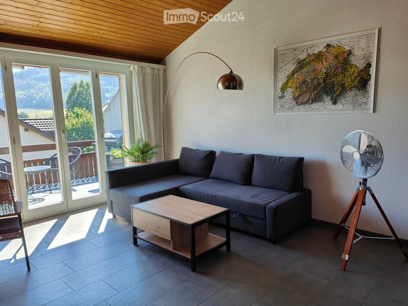 Modern living room with large windows offering a scenic view, featuring a grey sectional sofa, wooden coffee table, floor lamp, and a portable fan.