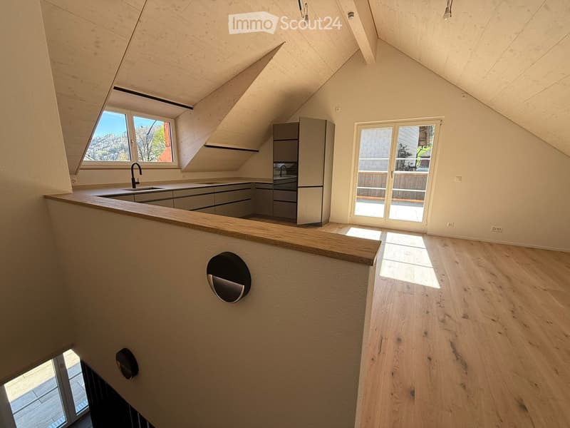 An empty attic space with wooden flooring, white walls, and large windows offering a view of the outdoors. The room features a modern kitchen area with cabinets and a sink, but no furniture or appliances are present.
