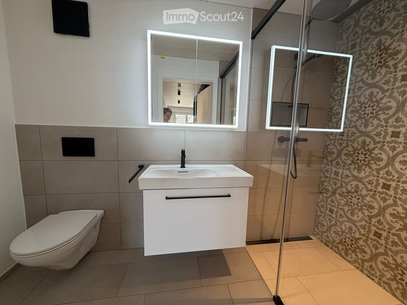 Modern bathroom with a white sink, toilet, illuminated mirrors, and a glass-enclosed shower area with decorative tiles.
