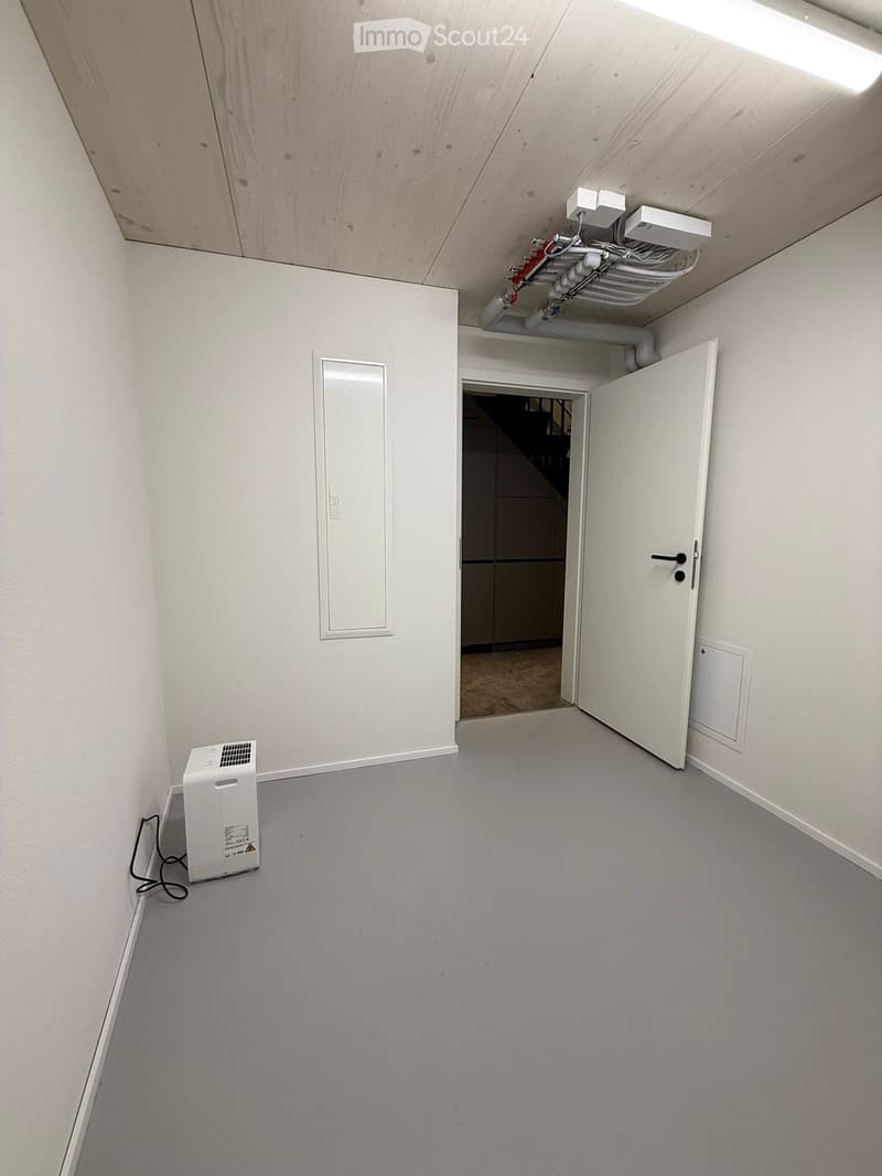 Empty room with grey carpeted floor, white walls, and a white door leading to another space. A small white appliance is placed near the left wall.