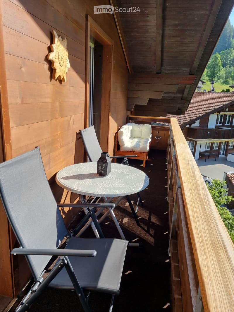A balcony with a wooden railing, a round table, and two chairs. A lantern is on the table. A cushion is on a chair. A wooden house is in the background.