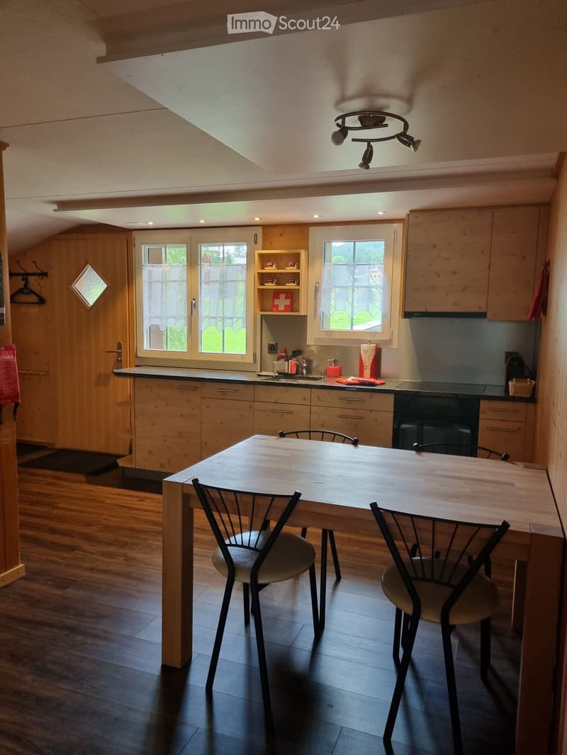 Wooden kitchen with dining table, chairs, cabinets, and a shelf with a red cross symbol. The kitchen has a sink, faucet, and a basket on the counter.