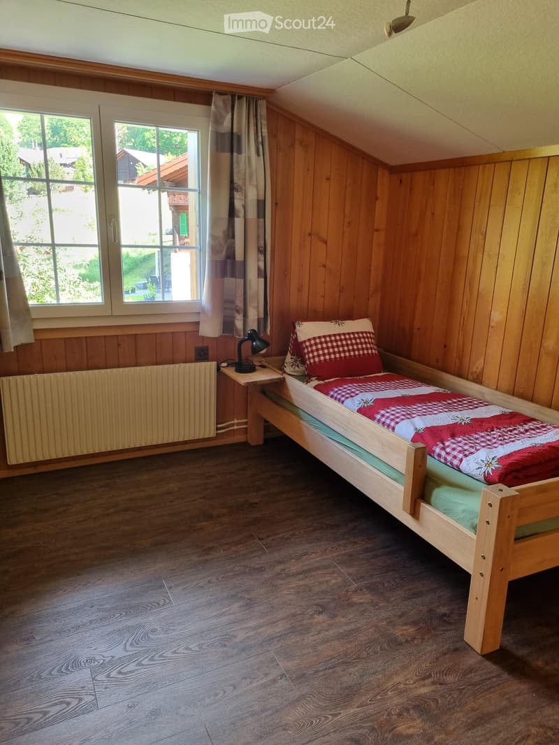 Small bedroom with wooden bed, red and white checkered bedding, pillow, lamp on nightstand, and radiator.