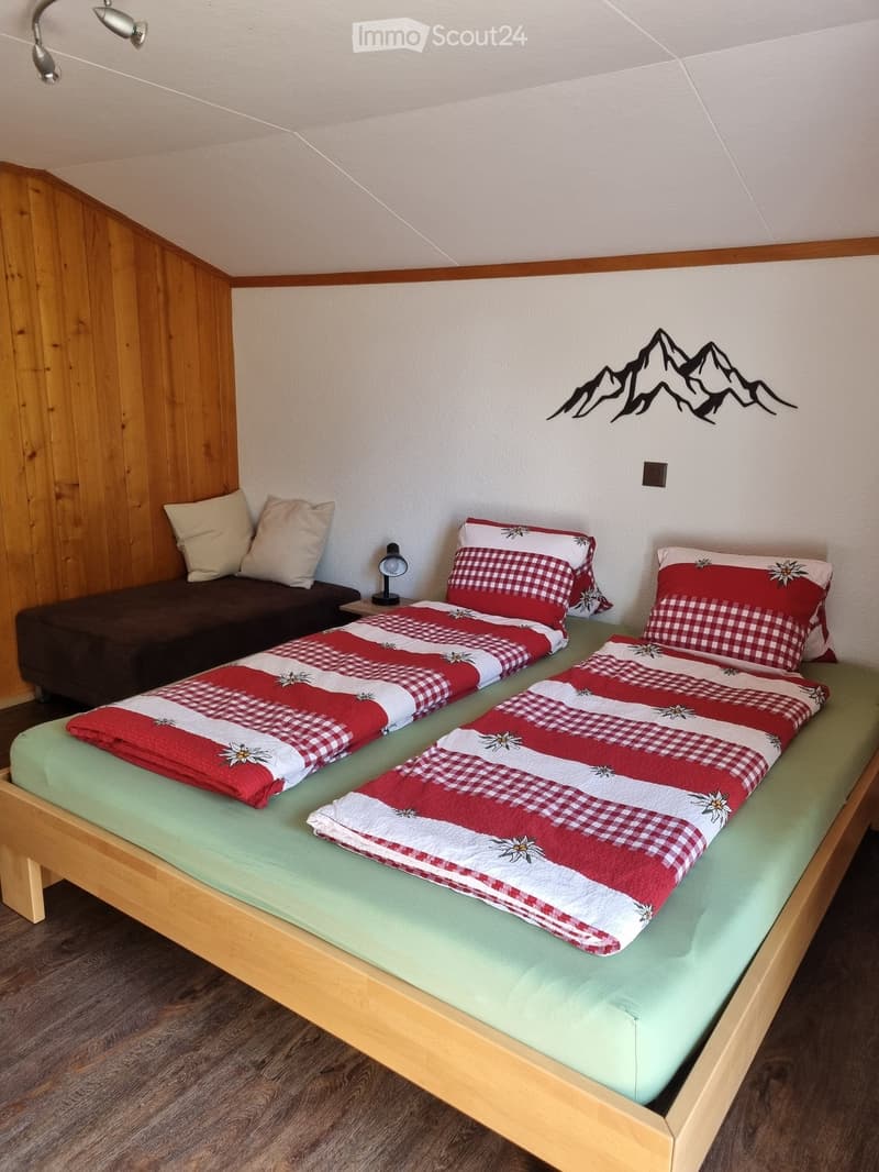 Bedroom with wooden bed, green mattress, red and white checkered bedspreads, pillows, nightstand with lamp, and a couch with pillows.
