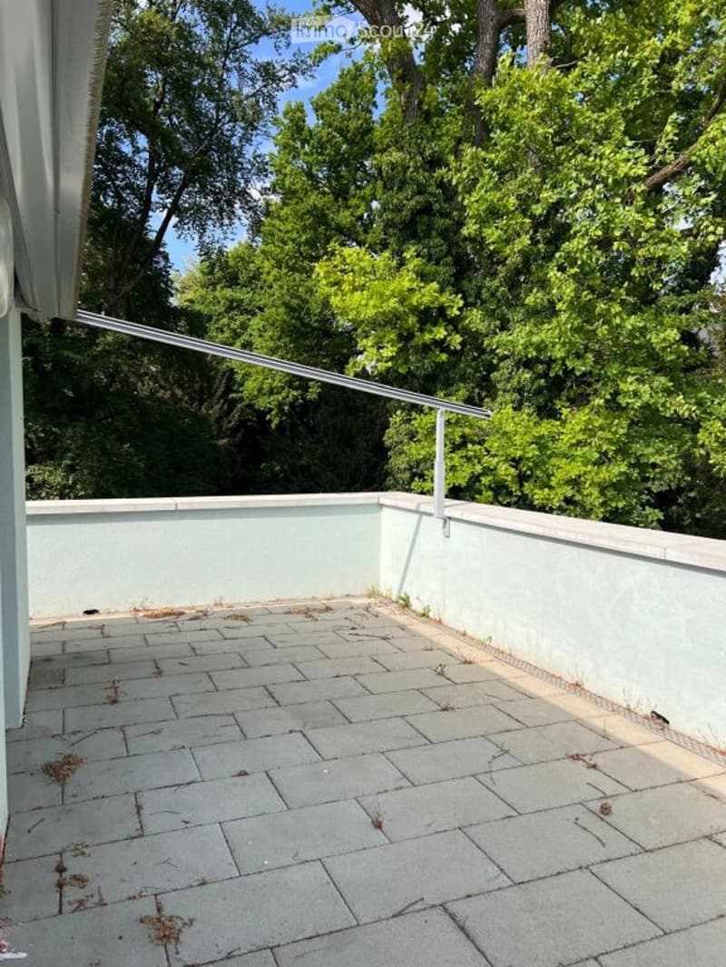 Empty balcony with light blue walls, tiled floor, metal railing, and lush green trees in the background.