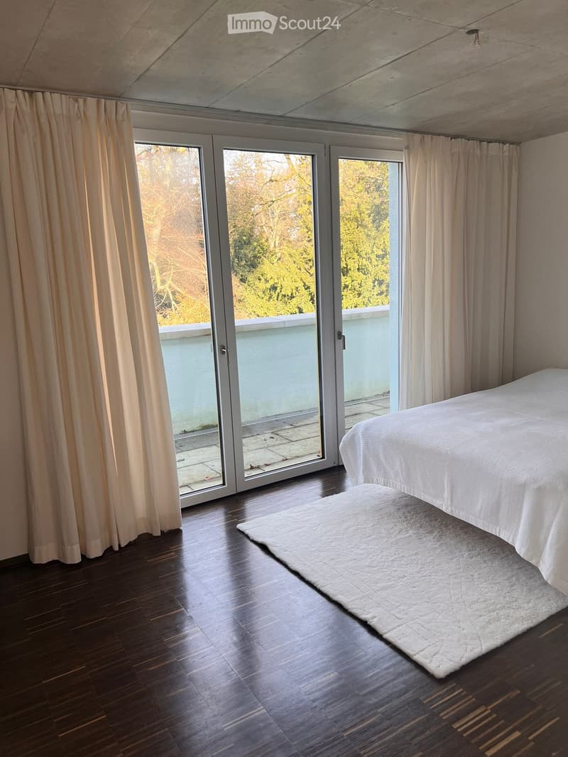Modern bedroom with wooden flooring, large sliding glass doors leading to a balcony, and a white bed with a shaggy rug.