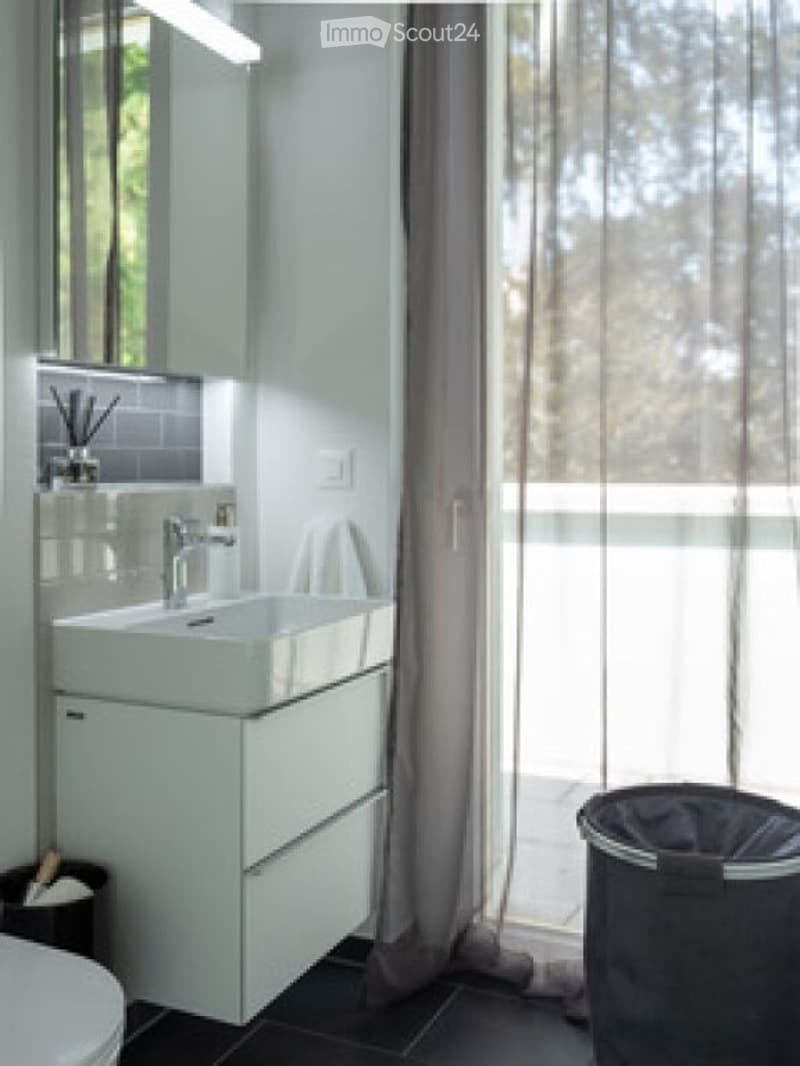Modern bathroom with a white sink, sleek faucet, large mirror, and a tall cabinet. The space features dark tiled flooring and a sheer curtain covering a window.