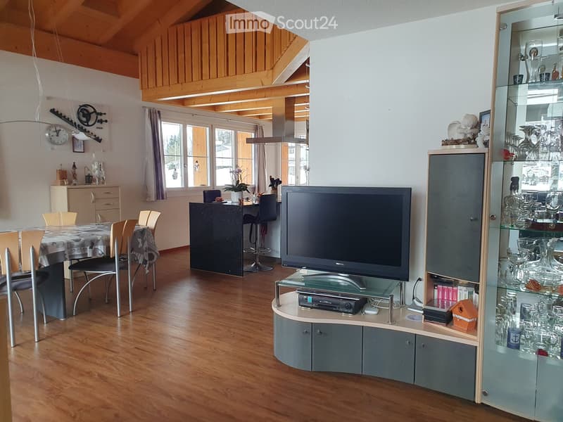 A spacious, well-lit living and dining area with wooden floors, a large television on a stand, a dining table with chairs, and a bar area with stools. The room features a mix of modern and traditional decor, including a glass cabinet displaying glassware