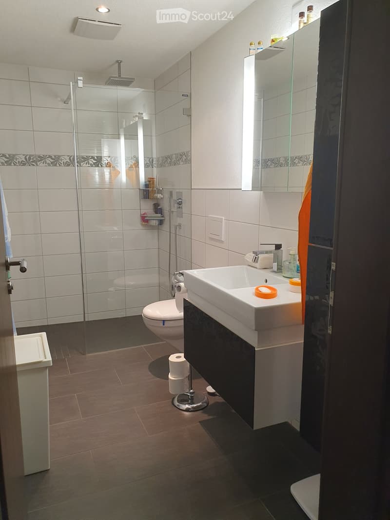 Modern bathroom with white tiles, a glass-enclosed shower, a toilet, a sink with a mirror, and a towel hanging on the side.