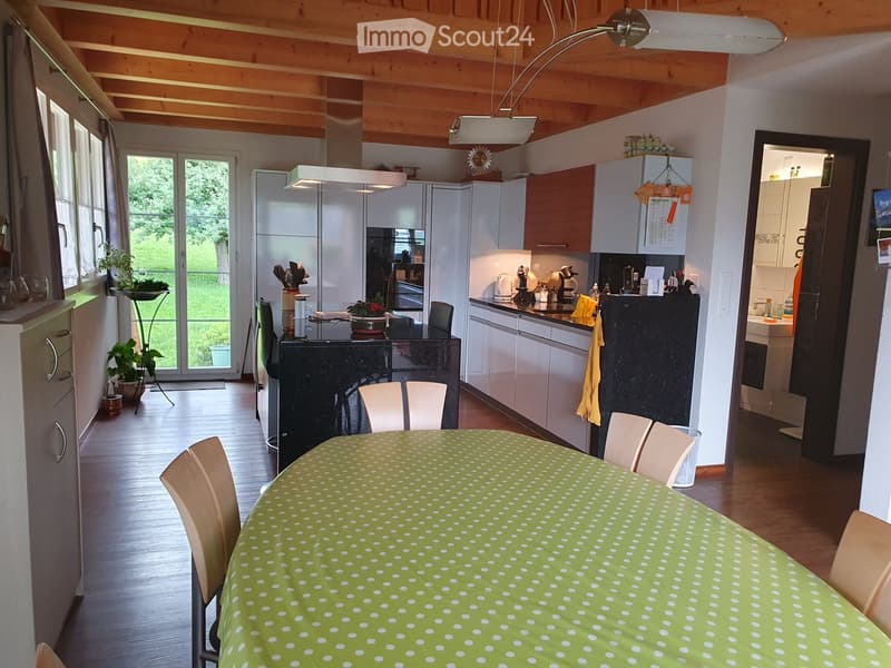 Open-plan kitchen and dining area with modern white cabinets, a black kitchen island, a dining table with chairs, and a refrigerator. The space features wooden flooring, large windows, and a view of greenery outside.