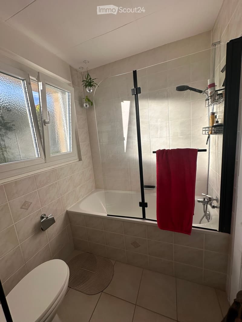 Modern bathroom with a bathtub, toilet, towel, and shower area. Features tiled walls and floor, a window with frosted glass, and a red towel hanging on a rack.