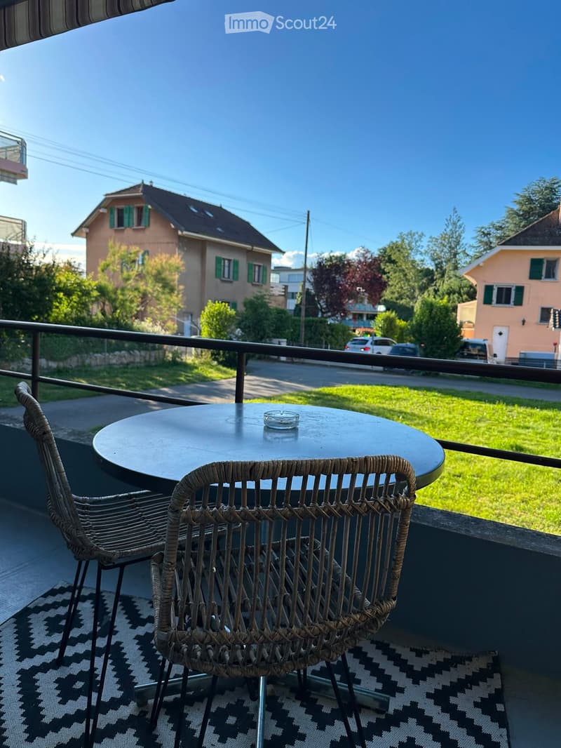 Balcony with a round table and two wicker chairs, overlooking a residential street with houses and greenery.