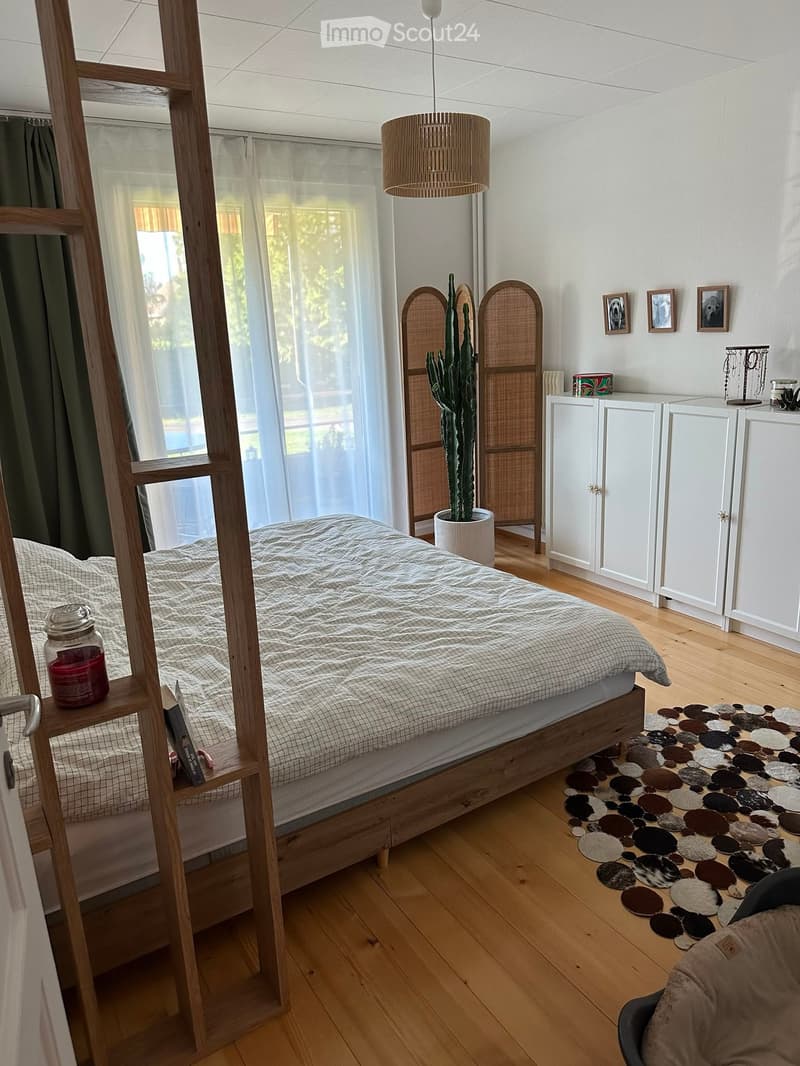 Bright bedroom with wooden floor, large window with sheer curtains, bed with grey bedding, white cabinets, cactus in a pot, and decorative items on shelves.
