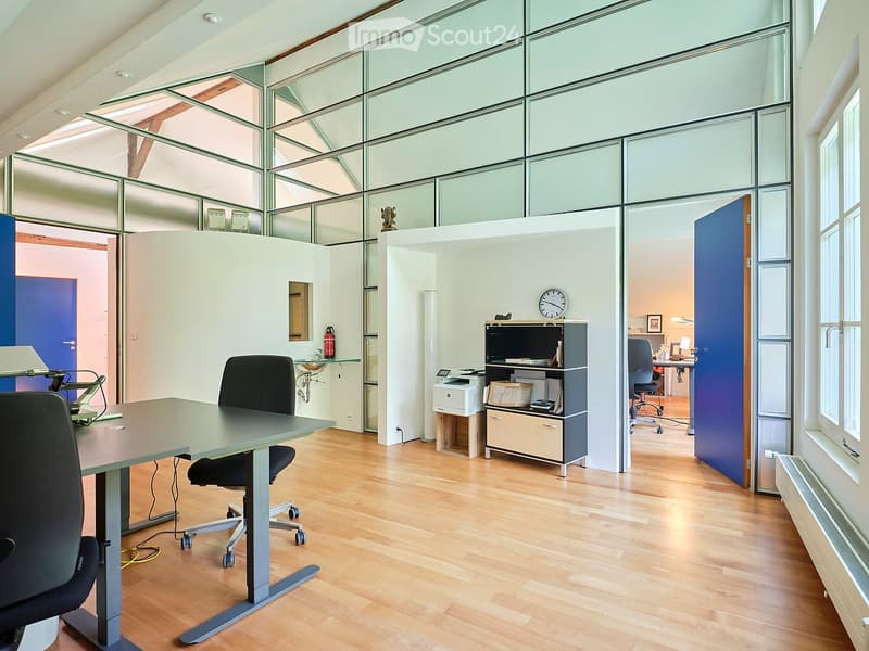 Modern office space with wooden flooring, large glass windows, a desk with chairs, a printer, a sink, and a clock on the wall.