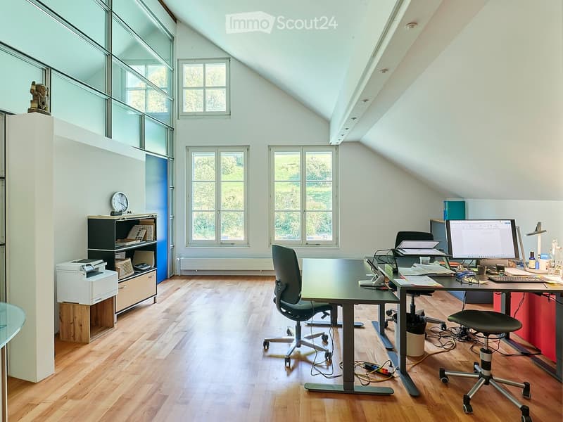 Bright, spacious attic office with wooden flooring, large windows, and modern furniture including a desk, chairs, computer setup, and shelving.