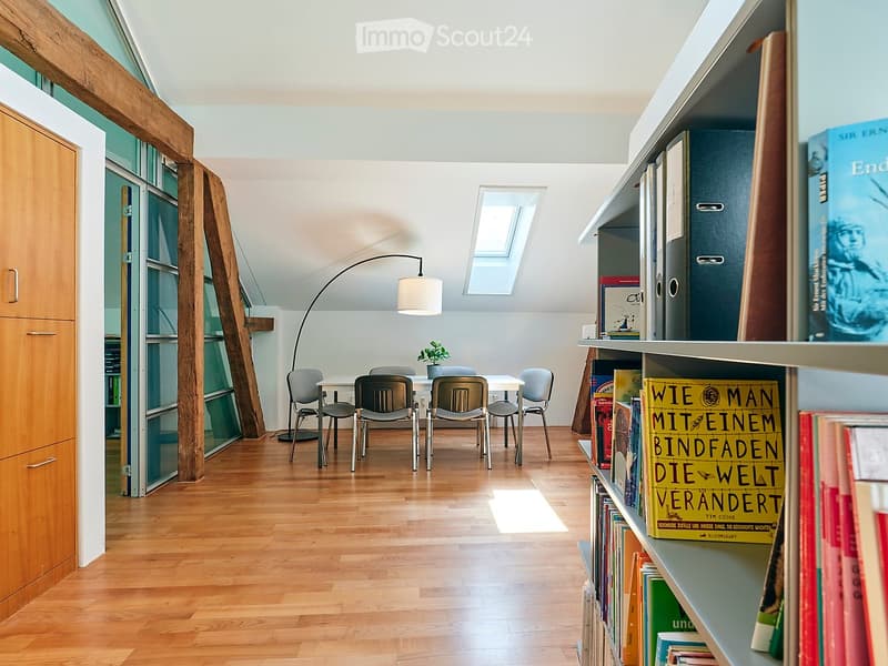 Bright, airy room with wooden flooring, large windows, and skylight. Features a dining table with four chairs, a bookshelf filled with books, a floor lamp, and a small plant on the table.