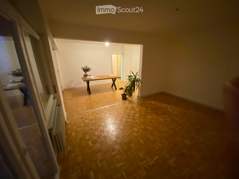 Empty room with wooden flooring and white walls, featuring a small table with a vase and a potted plant in the adjacent area.