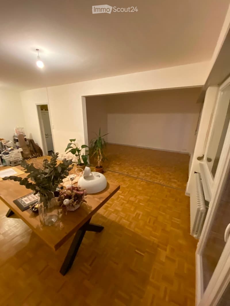 A wooden-floored room with a central wooden table holding a vase with flowers and a white radiator. The room has white walls and a visible doorway leading to another space.