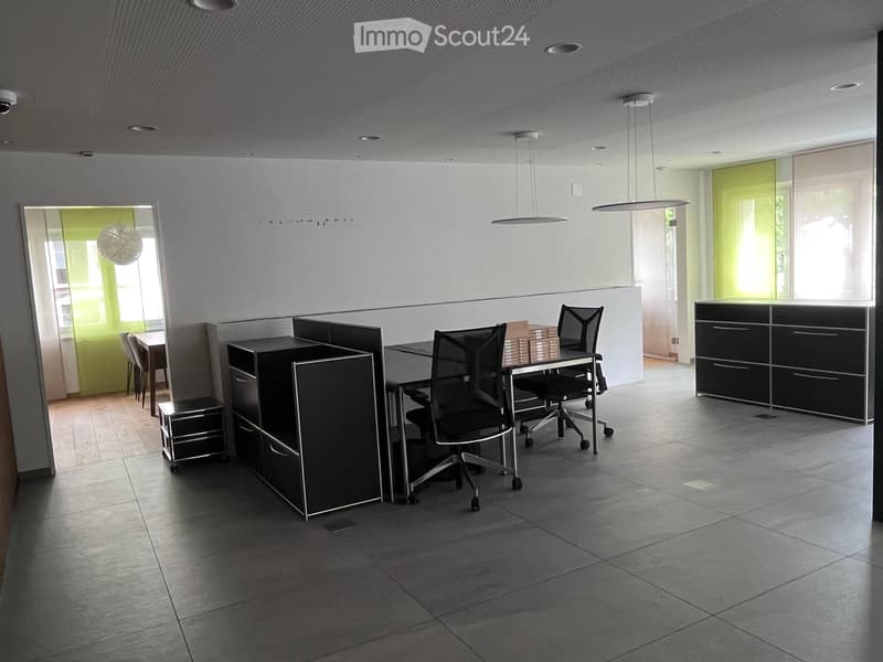 Modern office space with grey tiled floor, white walls, and large windows. Features include black office chairs, desks, cabinets, and a reception area with a counter.