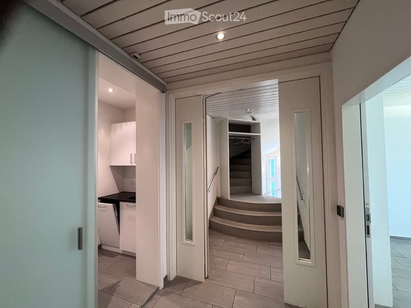 A modern hallway with a staircase leading upwards, featuring white walls, tiled flooring, and recessed lighting. A small closet and a doorway to another room are visible.