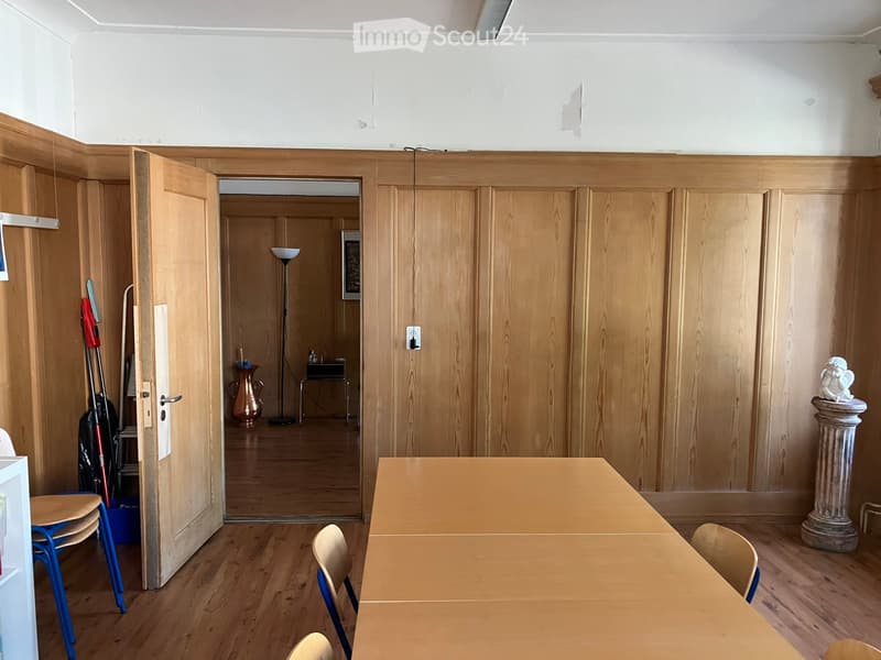 A furnished room with wooden panel walls, a large wooden table, several chairs, a floor lamp, a decorative vase, and a statue on a pedestal. The room has a wooden floor and an open doorway leading to another room.