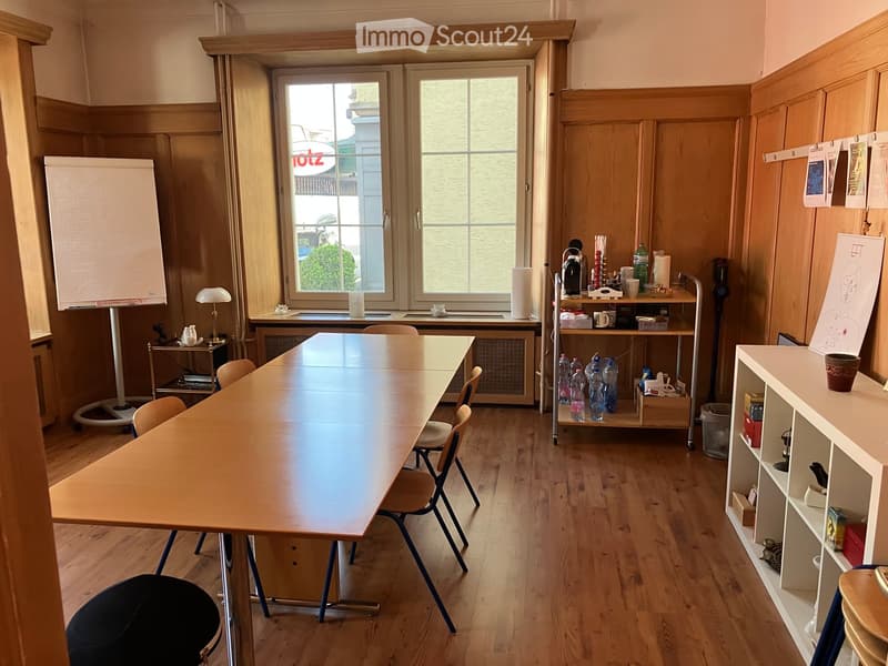 A furnished room with a large wooden table, chairs, a whiteboard, a lamp, and a small cabinet with various items on it.