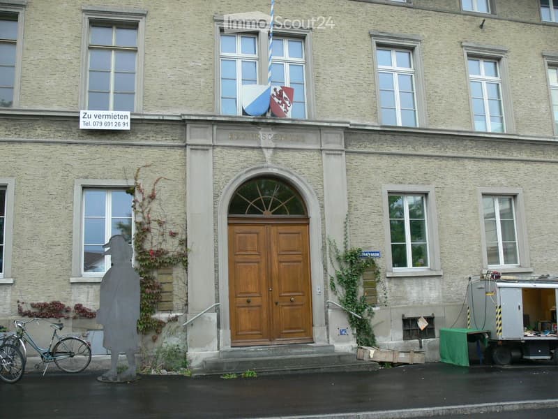 Facade of a multi-story building with large windows, a central wooden double door, and a flagpole with a flag and coat of arms above the entrance.
