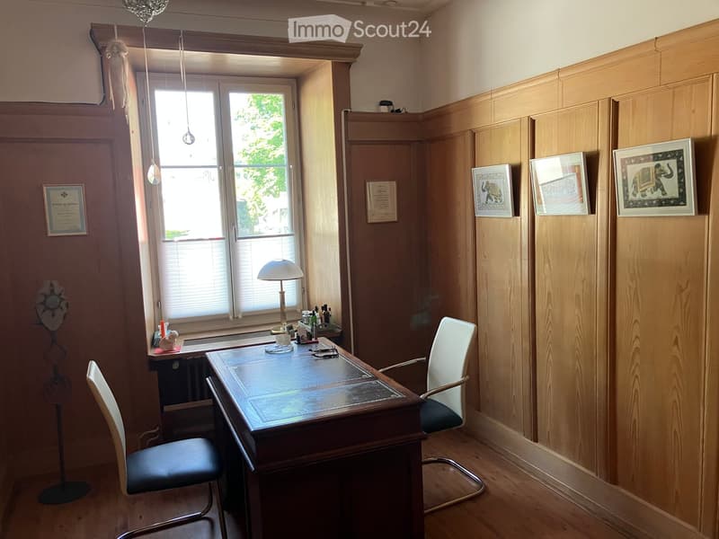 A well-lit room with wooden walls and flooring, featuring a large wooden desk with a glass top, two chairs, a table lamp, a standing fan, and several framed pictures on the wall.
