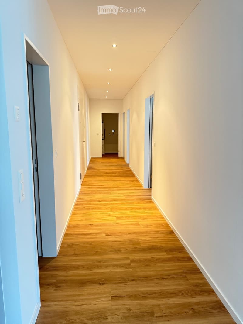 A clean, well-lit hallway with wooden flooring, white walls, and several doors leading to different rooms.